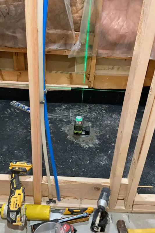Bathroom remodel in progress with exposed wall studs and insulation. A green laser level projects a vertical line, helping align the plumbing pipes and studs. Various construction tools, including a drill and utility knife, are scattered on the floor, indicating active work on the project.